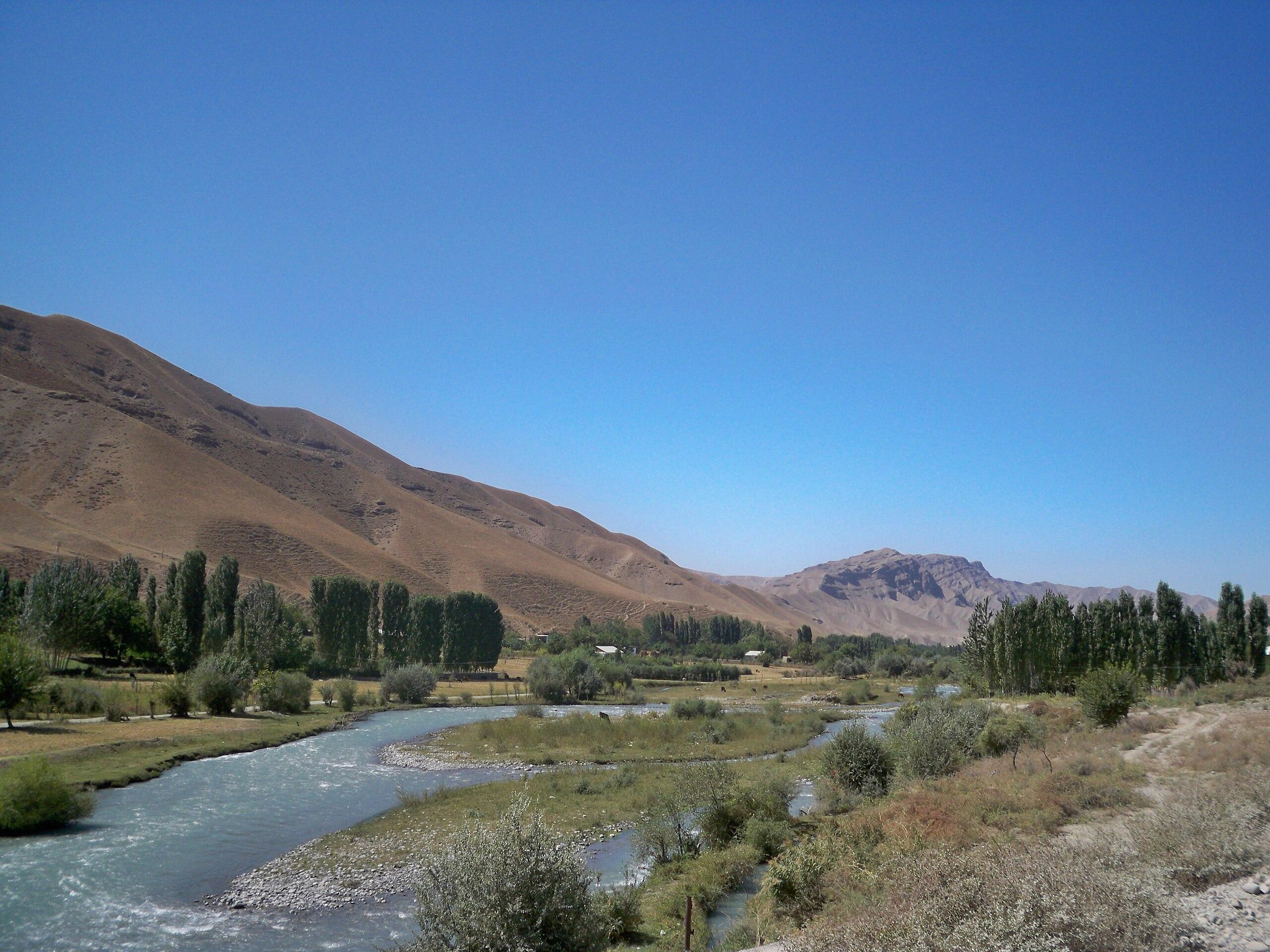 A view of the Khojabakirgan River flowing through the village of Layli in Leilek District, Kyrgyzstan. The river winds through a broad valley with scattered trees, farmland, and dry hills under a clear blue sky.